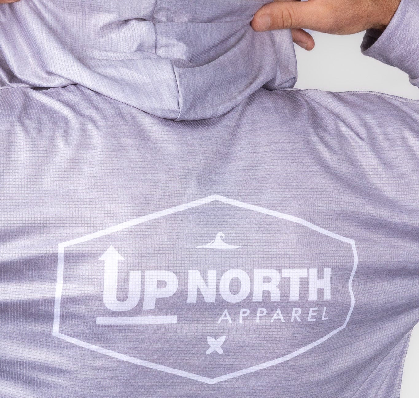 A man viewed from behind wearing a light lavender-purple hooded performance shirt with a white 'UP NORTH APPAREL' logo printed on the back, adjusting the hood.